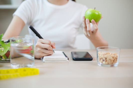 A person in a white t-shirt sits at a desk holding a green apple, is filling the habit tracker by the painless period clinic, with a glass of water, a smartphone, and a small bowl of nuts on the table.